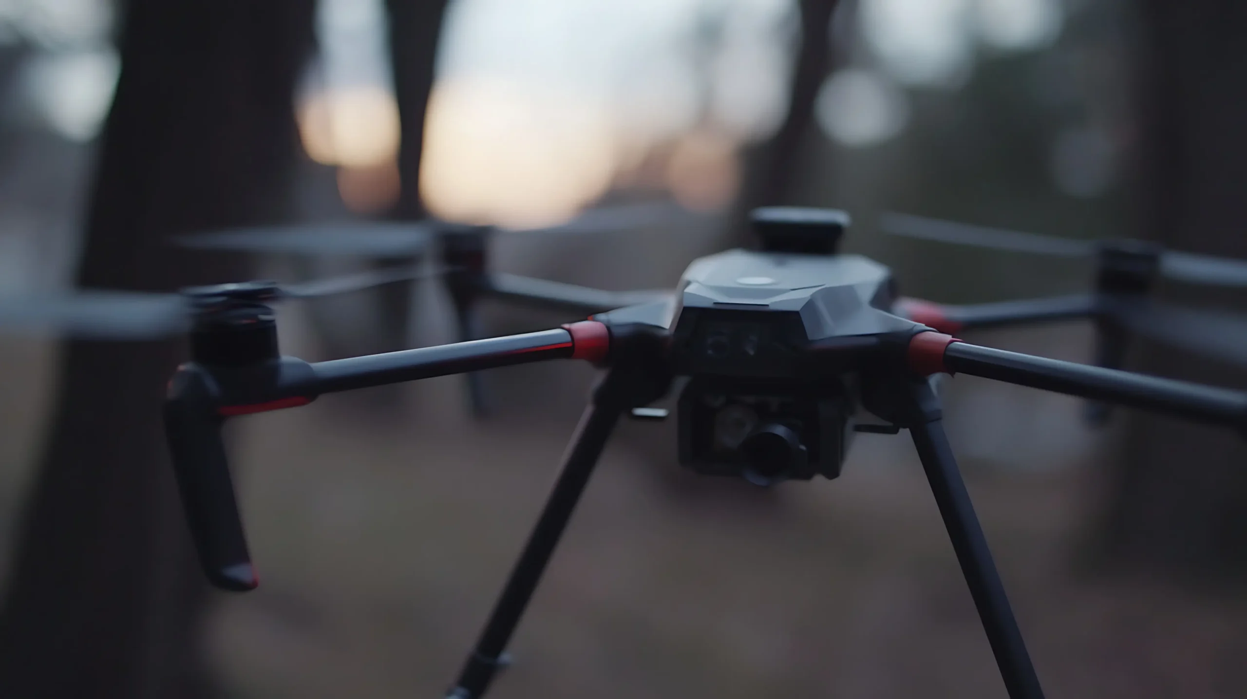 A close-up view of a black high-tech drone with a camera gimbal preparing for flight in a blurred outdoor wooded environment, showing its rotor arms and landing gear.
