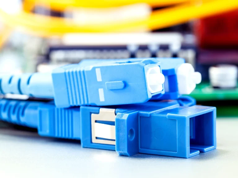 Close-up view of two blue fiber optic connectors, likely LC or SC duplex, showing the connector bodies and white ceramic ferrules, resting on a silver surface with a blurred green circuit board in the background.