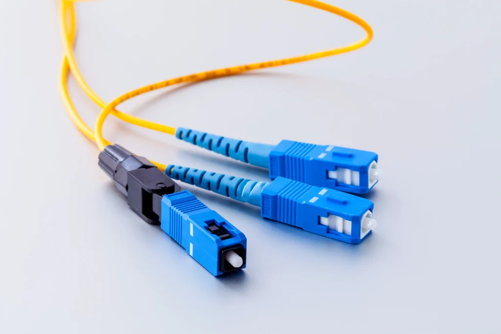 A close-up shot of two yellow fiber optic patch cables with various connectors on a light grey background. One cable has two blue LC UPC connectors. The other cable has a black LC connector and a blue LC UPC connector, showcasing different fiber optic connector types.