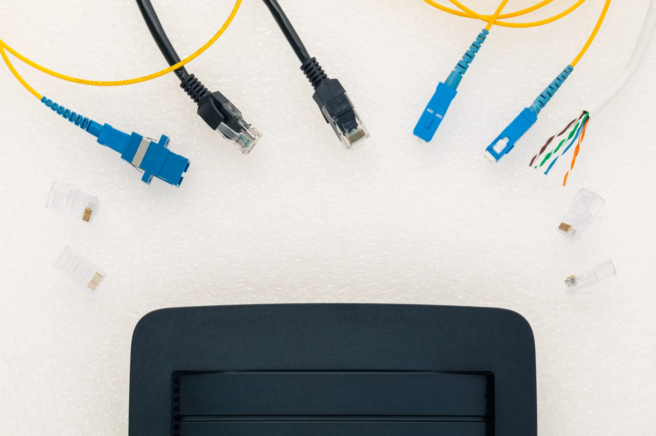 Overhead view of a black router and various network cables on a white surface, including blue fiber optic patch cords (LC and SC connectors), black Ethernet cables (RJ45), and loose wire pairs.